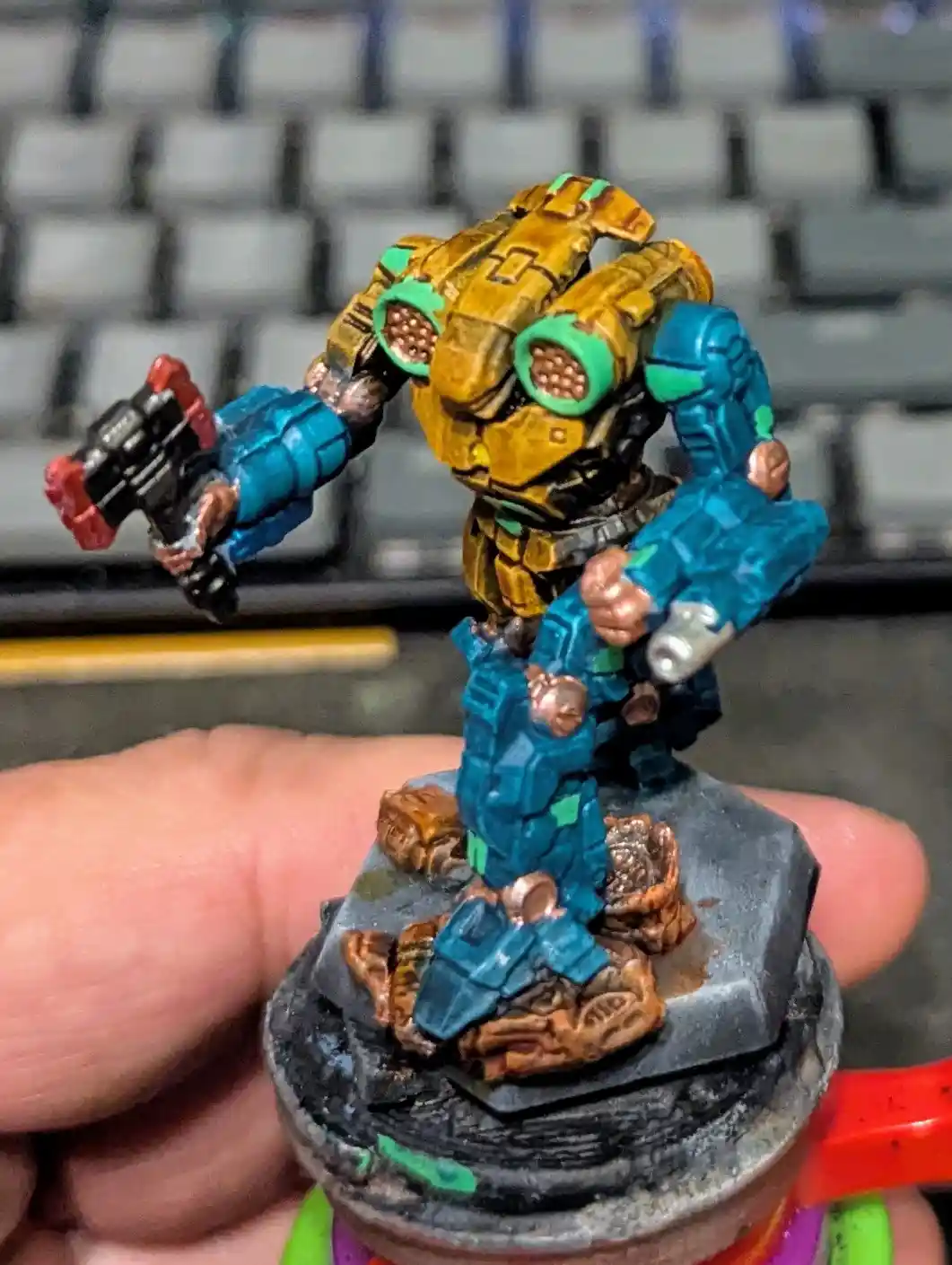 A hand holds a painted miniature of a blue and gold armored robot with a weapon, standing on a rocky base, in front of a…