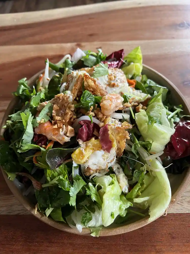 A close-up photo of a colorful salad in a wooden bowl, featuring mixed greens, cilantro, shredded carrots, bean sprouts…