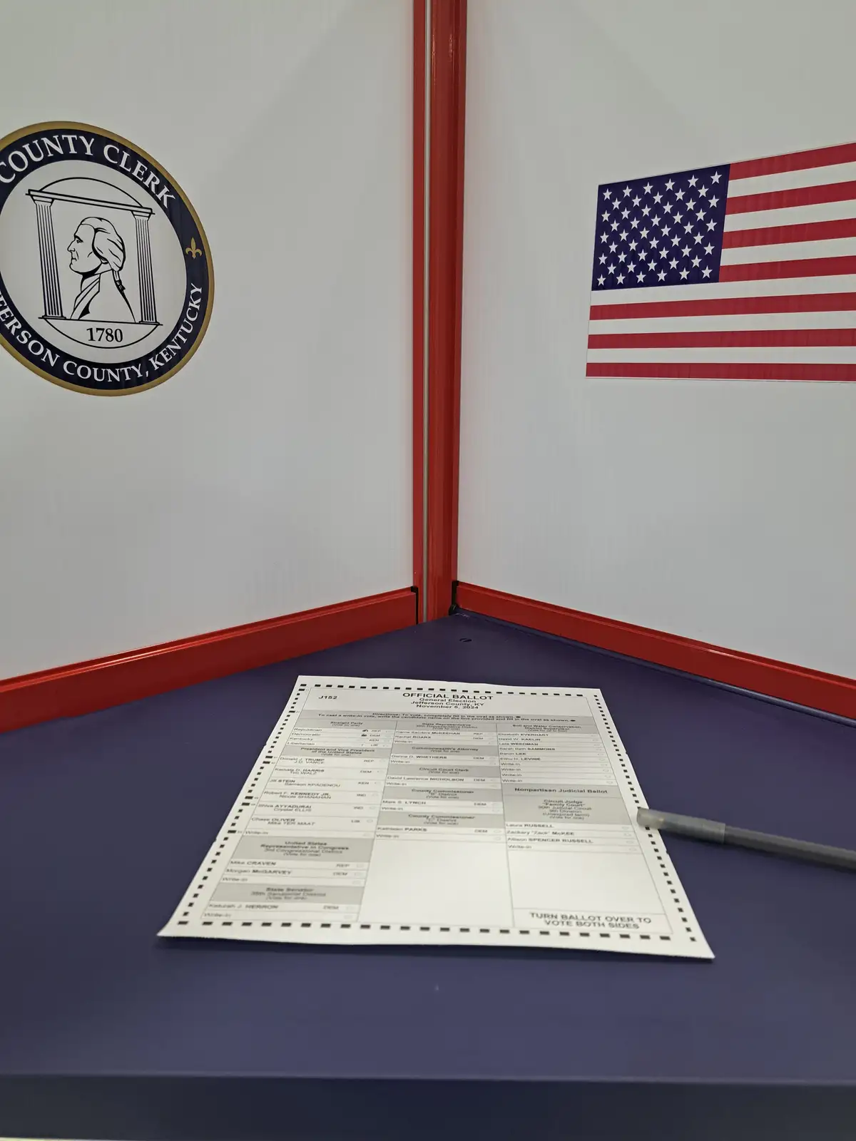 An official Kentucky election ballot lies on a blue voting booth surface, with a pen beside it. The booth walls show a…