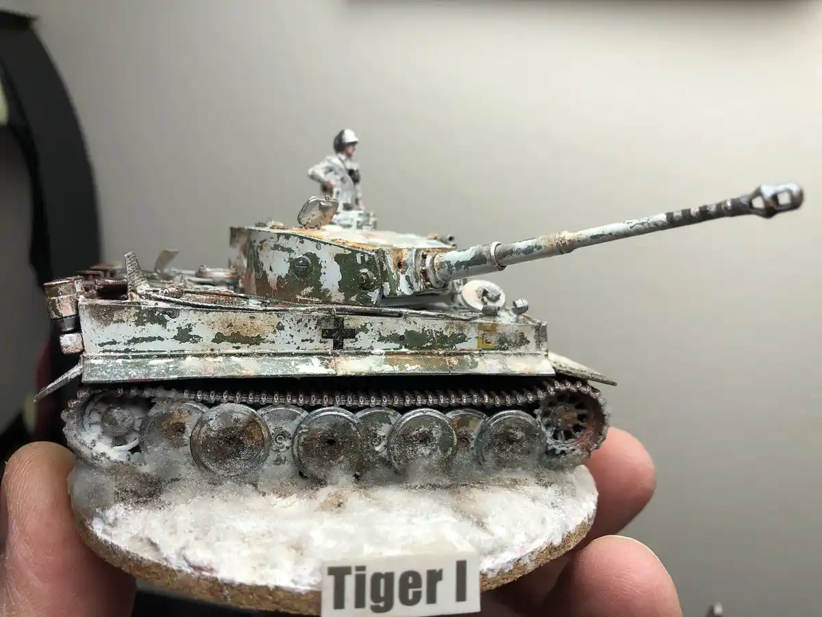 A hand holds a highly detailed, weathered model of a Tiger I tank with a miniature soldier figure on top. The tank's paint…