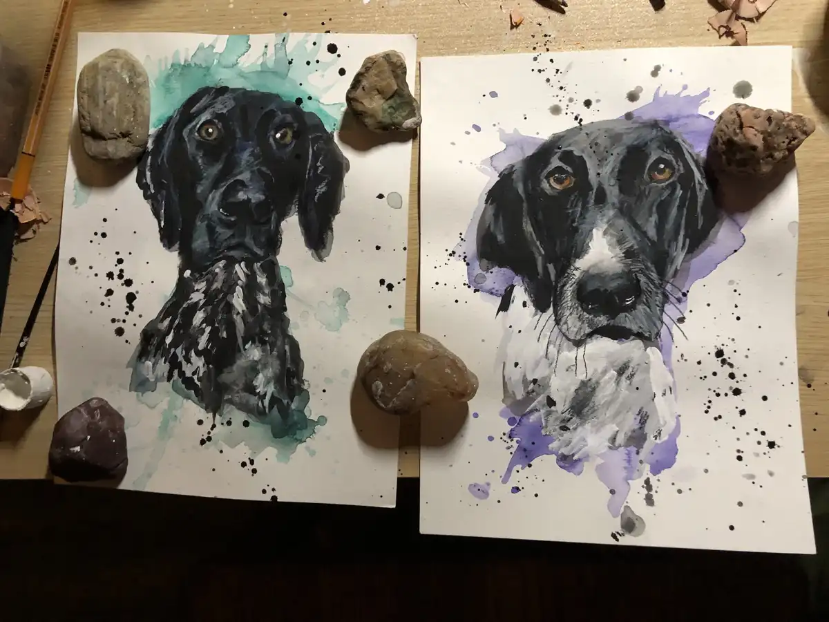 Two watercolor dog portraits sit side-by-side on a wooden table, each surrounded by real rocks placed as if they were part…