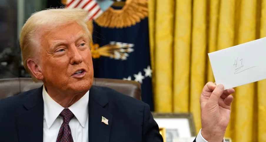 Former president Donald Trump is seated in the Oval Office, holding up a small white card with the handwritten number '47'…