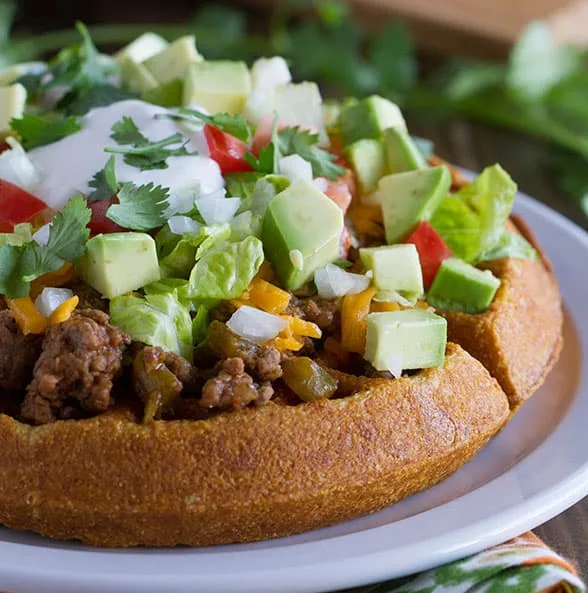 A close-up photo of a loaded taco bowl made from a round, baked bread base, filled with seasoned ground meat, cheese, diced…