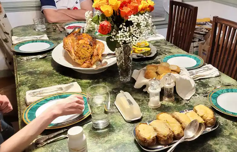 A dining table is set for a formal meal with a roasted turkey, cheesy baked potatoes, rolls, and a floral centerpiece, but a…
