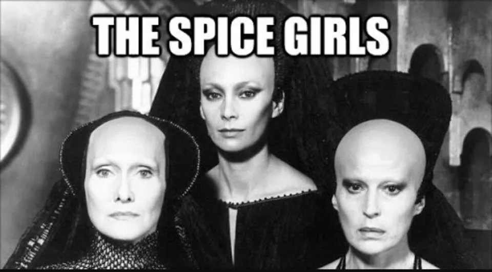 A black-and-white image shows three women with shaved heads and dark, high-collared costumes, resembling characters from the…