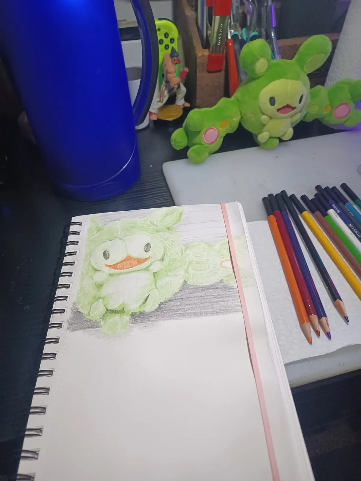 A spiral-bound sketchbook lies open on a desk, displaying a colored pencil drawing of a green, fluffy, caterpillar-like…