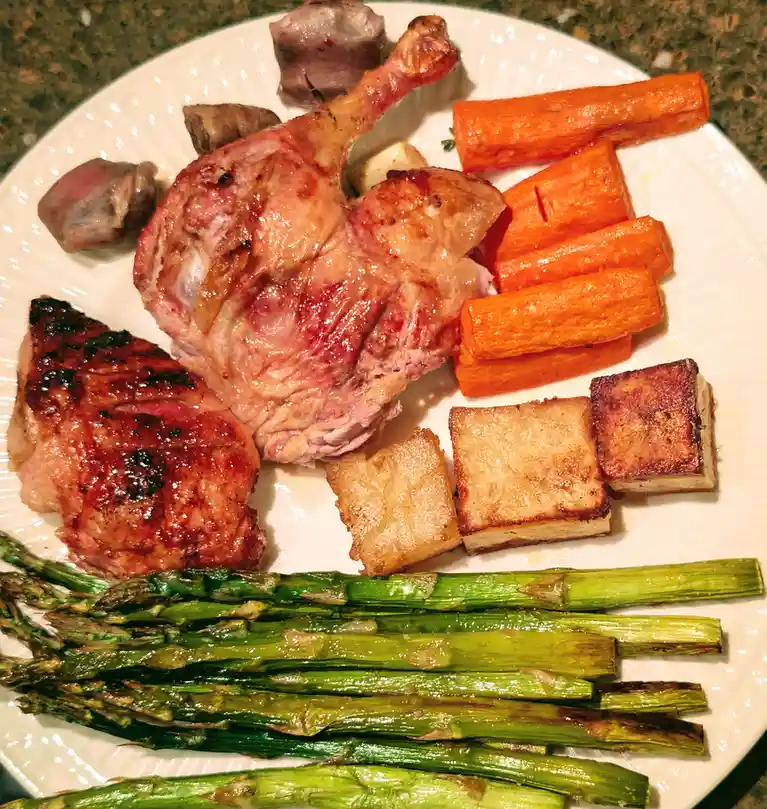 A white plate holds a generous, visually appealing meal featuring roasted duck leg, grilled duck breast, roasted carrots…