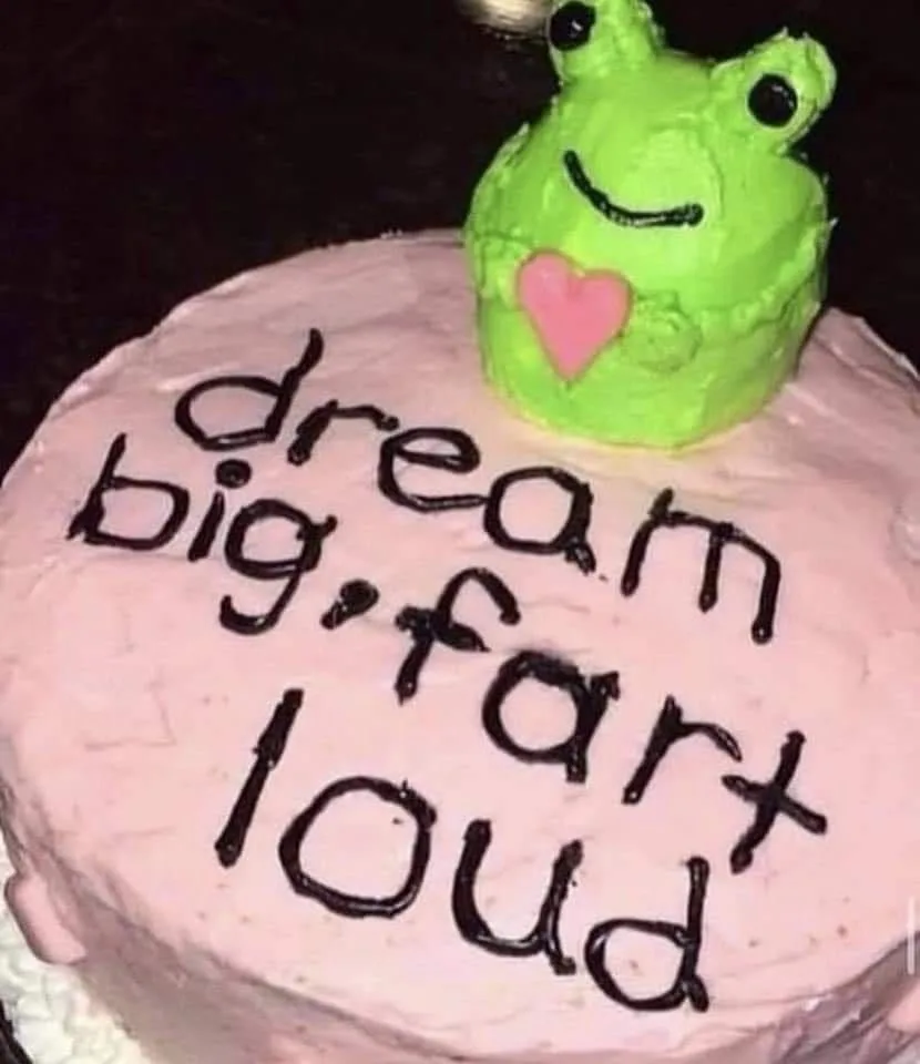 A pink frosted cake has the phrase 'dream big, fart loud' written in black icing. On top sits a green, smiling frog-shaped…