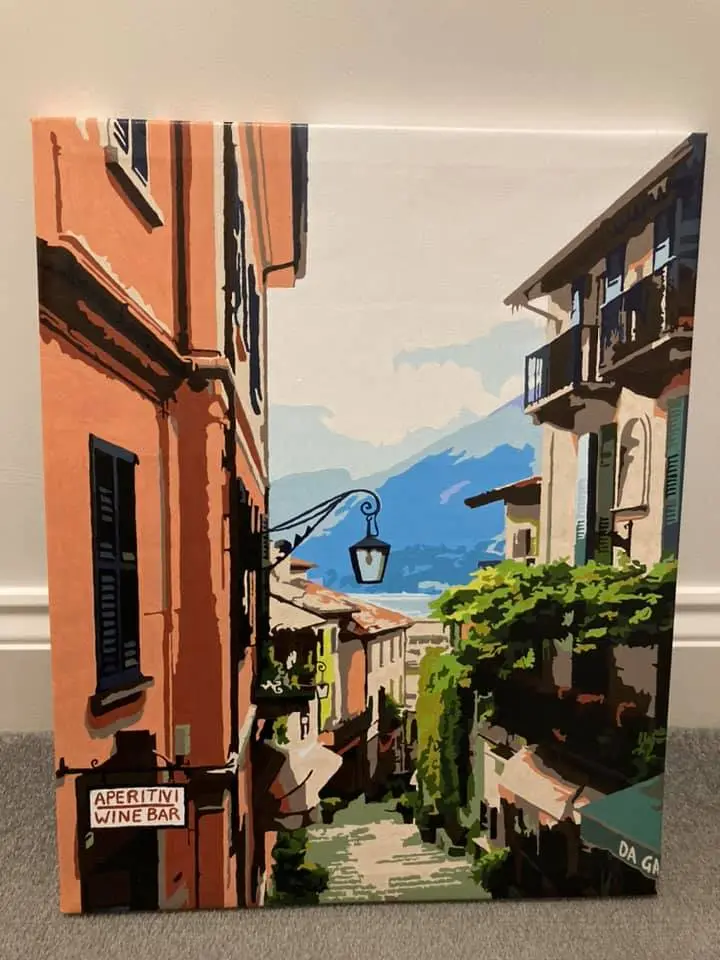 A painting of a picturesque European street with orange buildings, greenery, and mountains in the background. A sign on the…