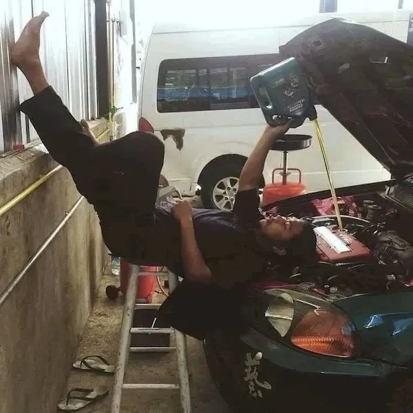 A person is lying horizontally on a ladder, one leg kicked up against a wall, pouring motor oil into a car's engine while…