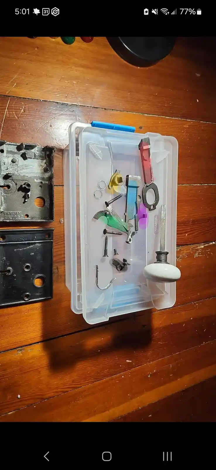 A clear plastic toolbox filled with assorted small tools and hardware sits open on a wooden floor, next to two disassembled…