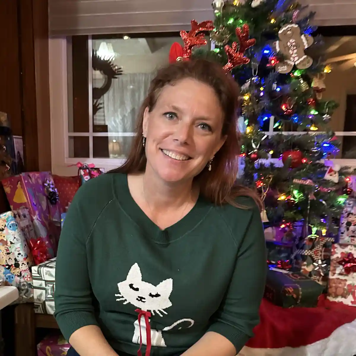 A woman with reddish-brown hair smiles while wearing red reindeer antlers and a green sweater with a white cat design…