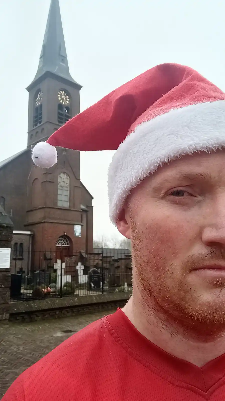 A man wearing a red and white Santa hat and a red shirt takes a selfie in front of a brick church with a clock tower. The…