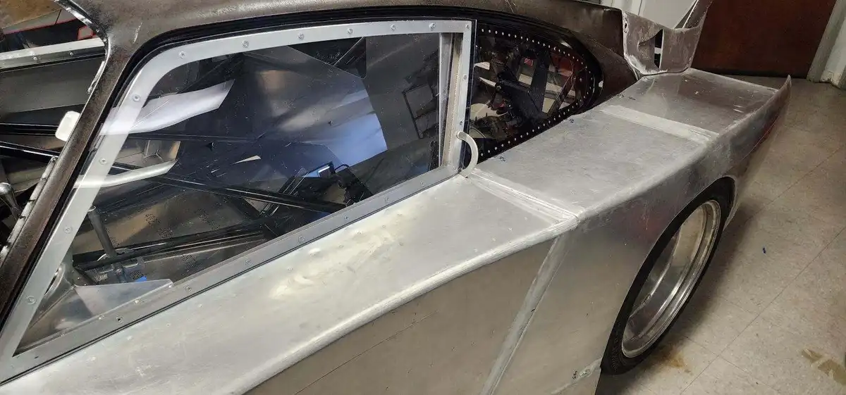 A close-up of a silver, stripped-down race car with a partially installed window frame, showing the bare metal body and…