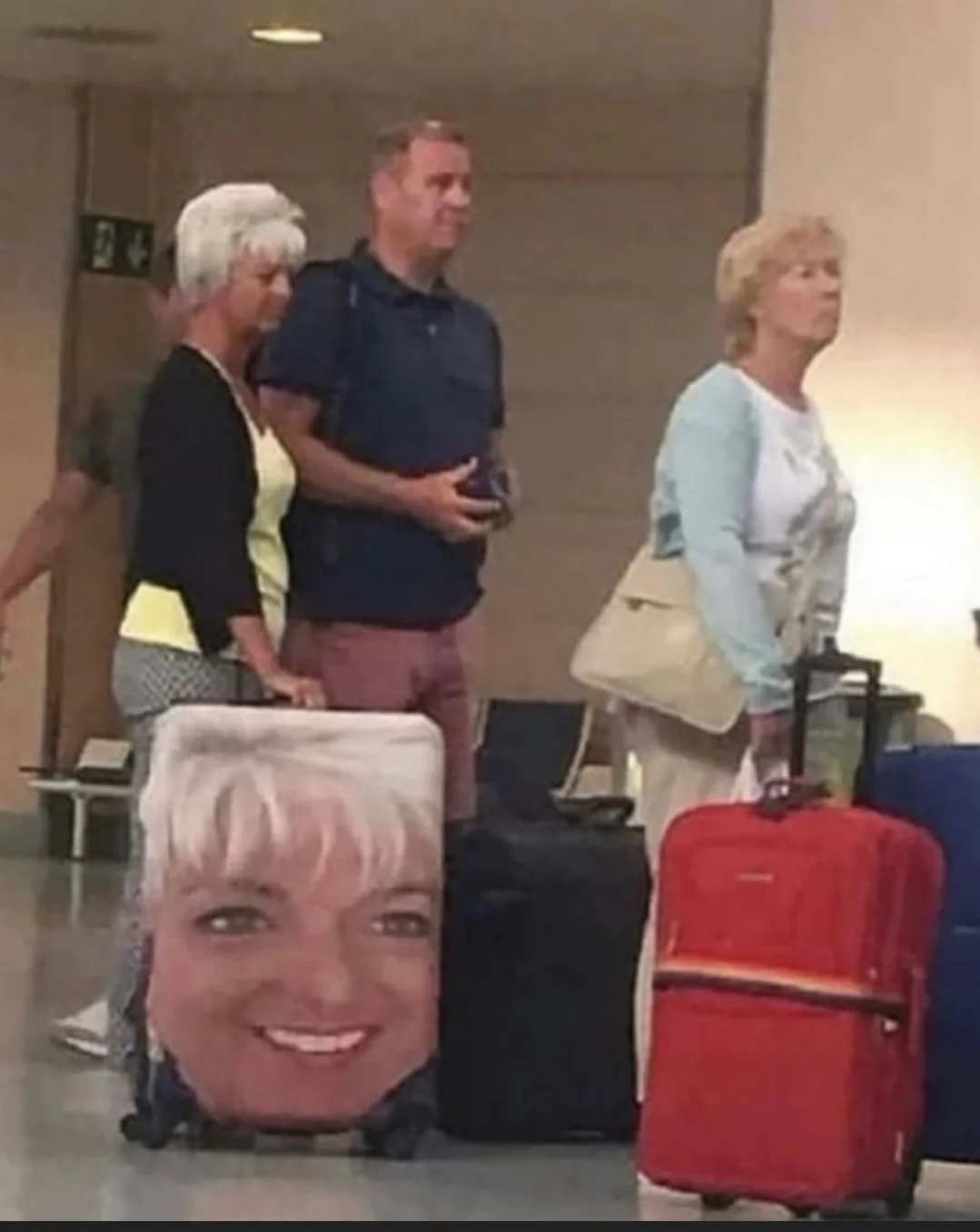 Three people stand in an airport terminal with luggage; one woman's suitcase has a large printed photo of her own smiling…