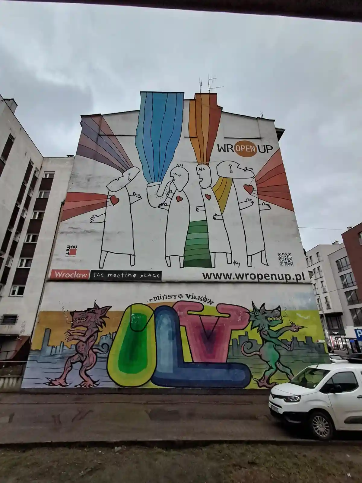 A large mural on a building shows four stylized figures in white robes with colorful beams emanating from their heads, one…