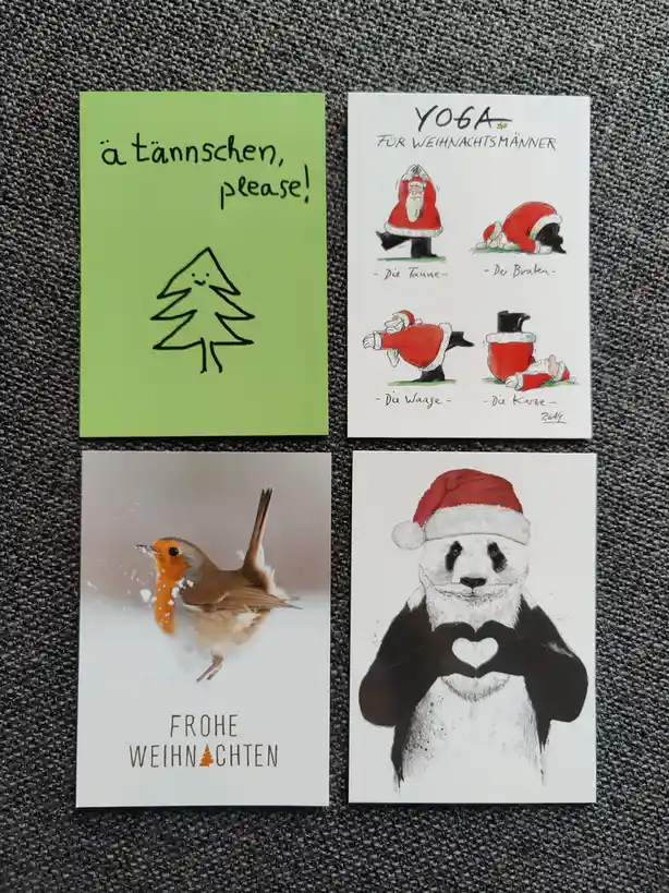 Four Christmas cards arranged on a gray fabric surface. Top left: a green card with a simple black line drawing of a smiling…