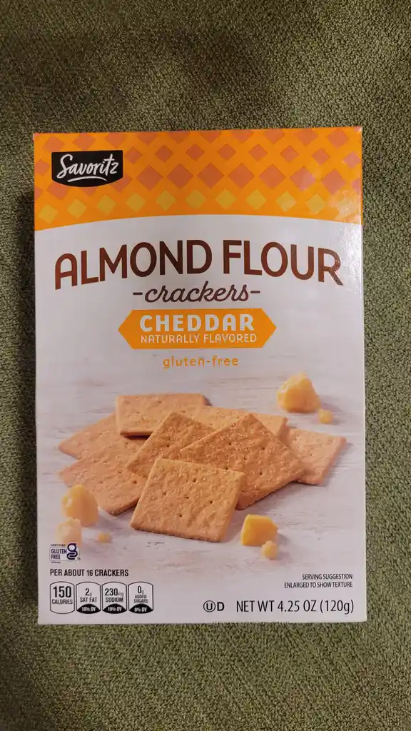 A box of Savoritz brand almond flour crackers, cheddar flavored and gluten-free, sits on a green textured surface. The…
