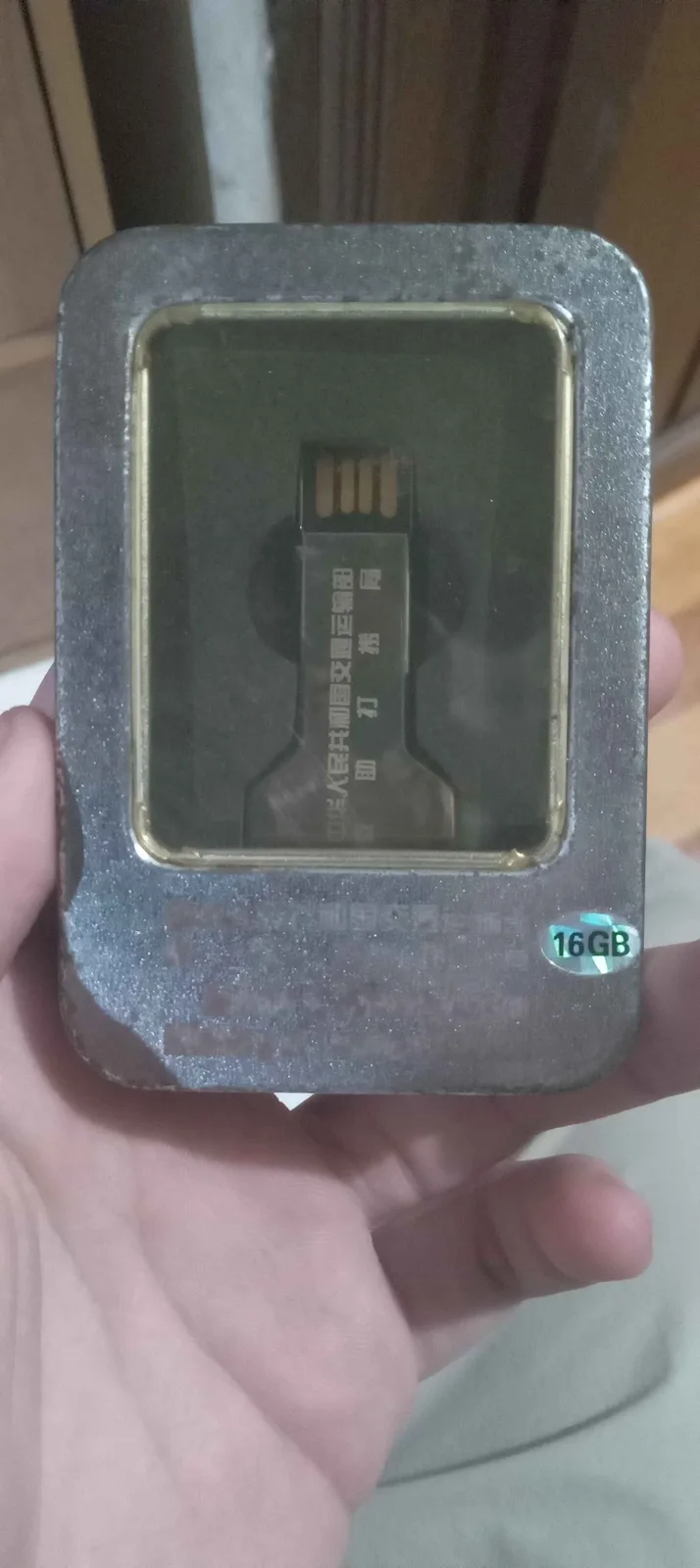A hand holds a gray, rectangular, metallic-looking device with a transparent window revealing a USB flash drive inside. The…