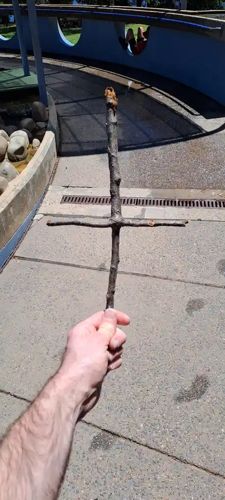 A person's hand holds a stick that has been broken and arranged into a crude cross shape, standing upright on a paved…