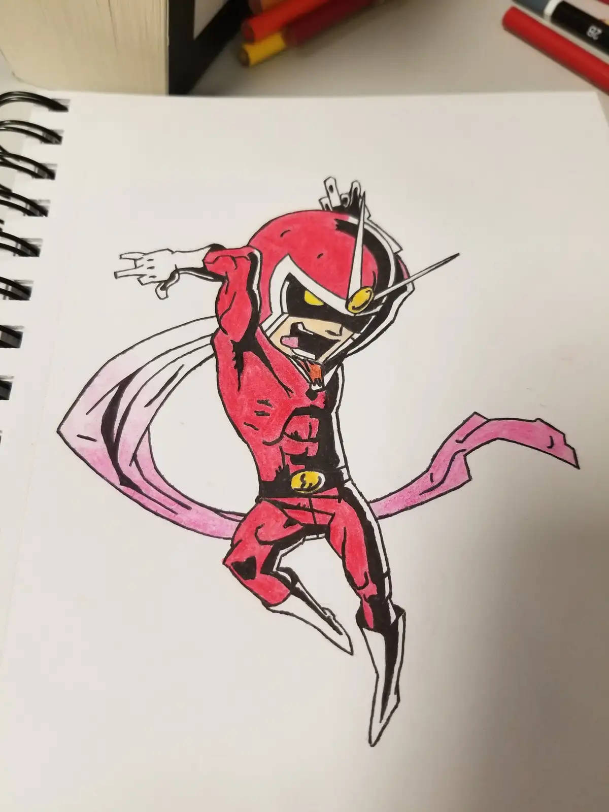 A hand-drawn cartoon superhero in a red suit with a pink cape and helmet, drawn on a spiral-bound notebook. The character…