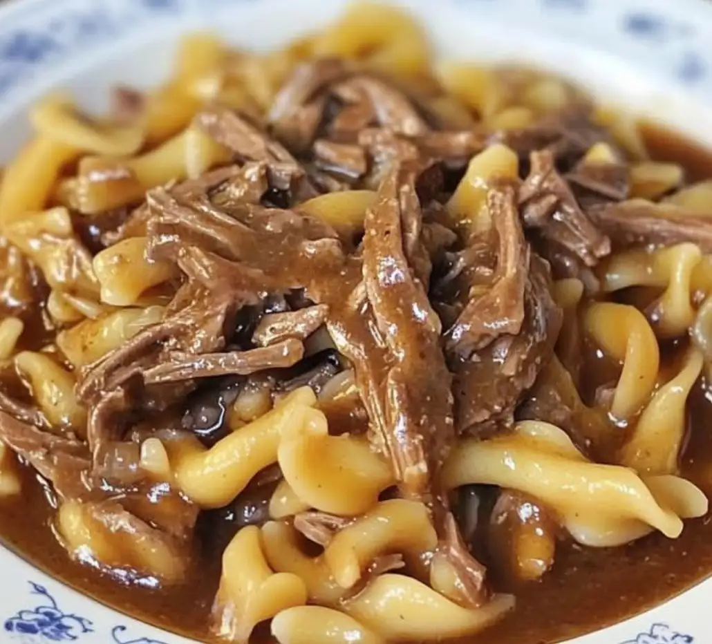 a close-up photo of a bowl of pasta covered in shredded meat and brown gravy, served on a blue and white patterned plate…