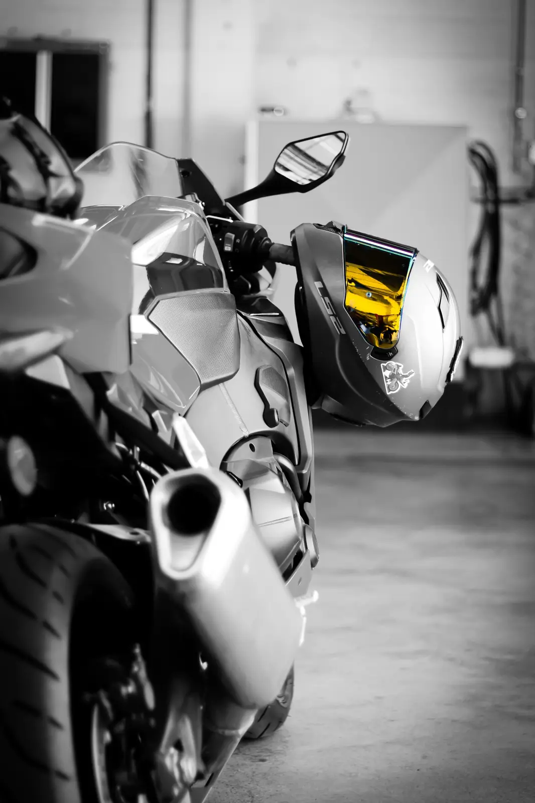 A black-and-white photo of a sport motorcycle in a garage, with a helmet hanging from the handlebars. The helmet's visor is…