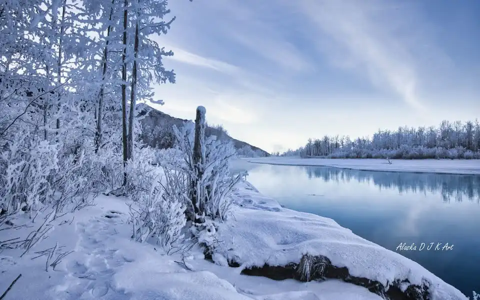 A serene winter landscape photograph showing snow-covered trees and bushes lining a frozen river under a pale blue sky with…