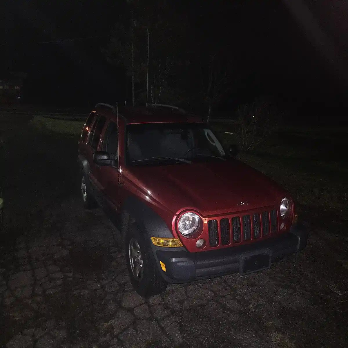A red Jeep Liberty is parked on a dark, cracked asphalt surface at night, with trees and darkness in the background. The…