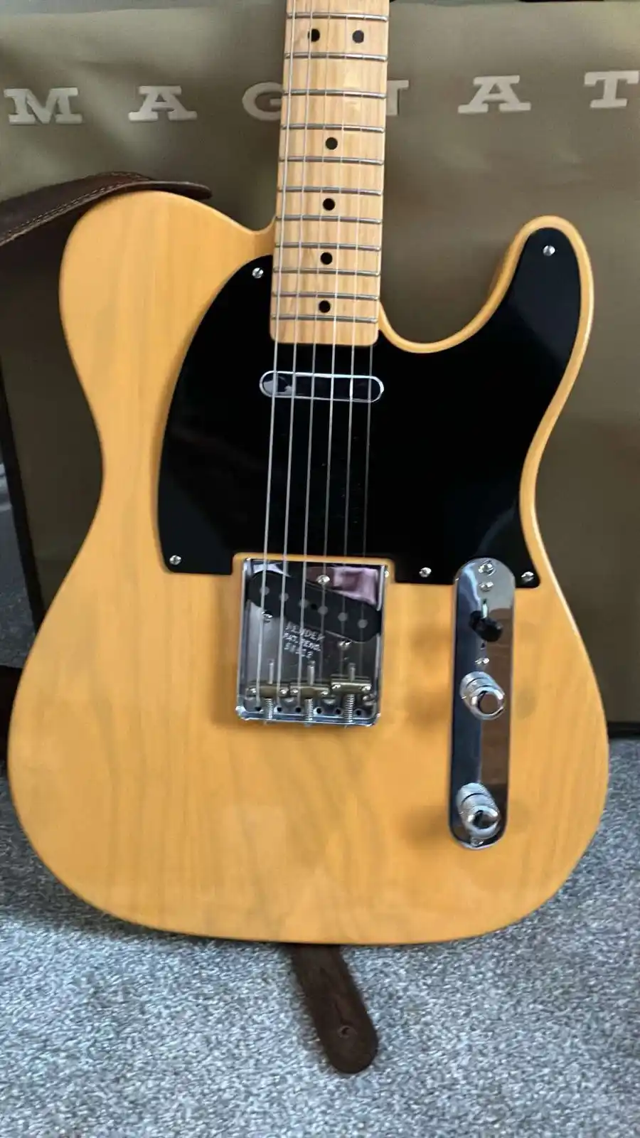 A light-wood Fender Telecaster-style electric guitar with a black pickguard and chrome hardware is shown upright, resting on…