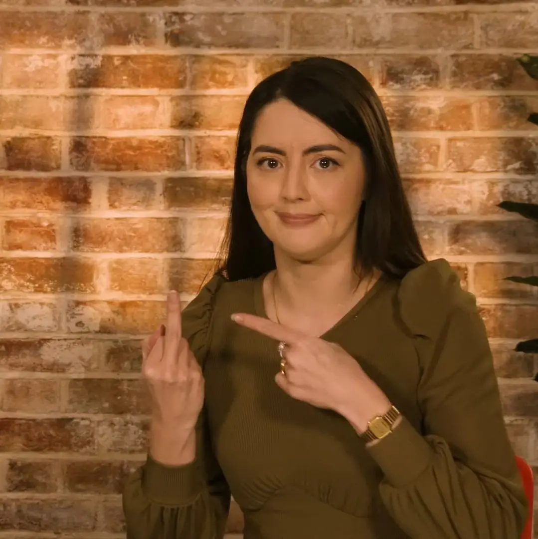 a woman with dark hair and a green top sits in front of a brick wall, smiling while pointing at her raised middle finger…