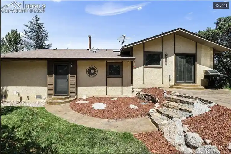 a single-story beige house with a brown roof, a front door with wooden siding, and a backyard featuring a circular path made…