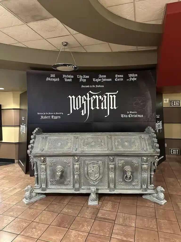 A large, ornate, silver-colored coffin with skull and lion-head decorations sits on a tiled floor in front of a movie poster…