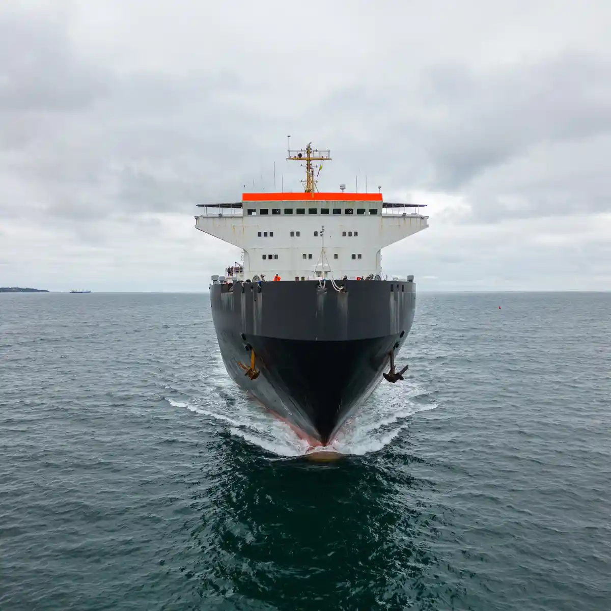 A large cargo ship with a white superstructure and orange roof sails directly toward the viewer on a cloudy day, its bow…