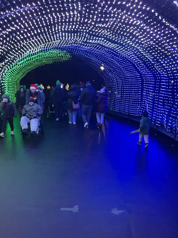 A group of people, including a man in a wheelchair and a child with a toy, walk through a brightly lit tunnel adorned with…