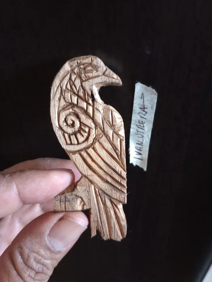 A hand holds a carved wooden bird with intricate geometric patterns, next to a torn label that reads 'IVANUTAERA' pointing…