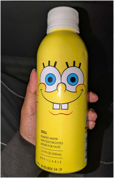 A hand holds a yellow reusable water bottle featuring a large, cheerful cartoon face resembling SpongeBob SquarePants. The…