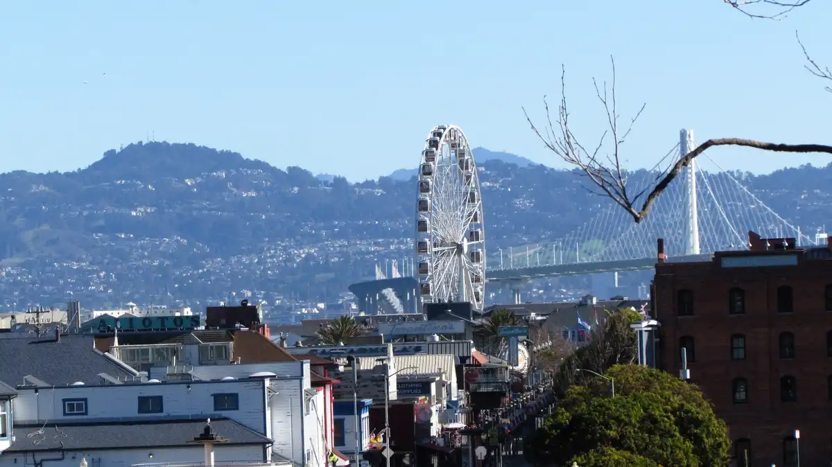 A scenic daytime photo of a cityscape featuring a large white Ferris wheel, a modern suspension bridge, and rolling hills in…