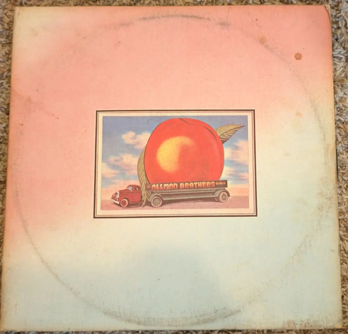 A vintage, worn record album cover with a faded pink and cream gradient background. The central artwork depicts a red…