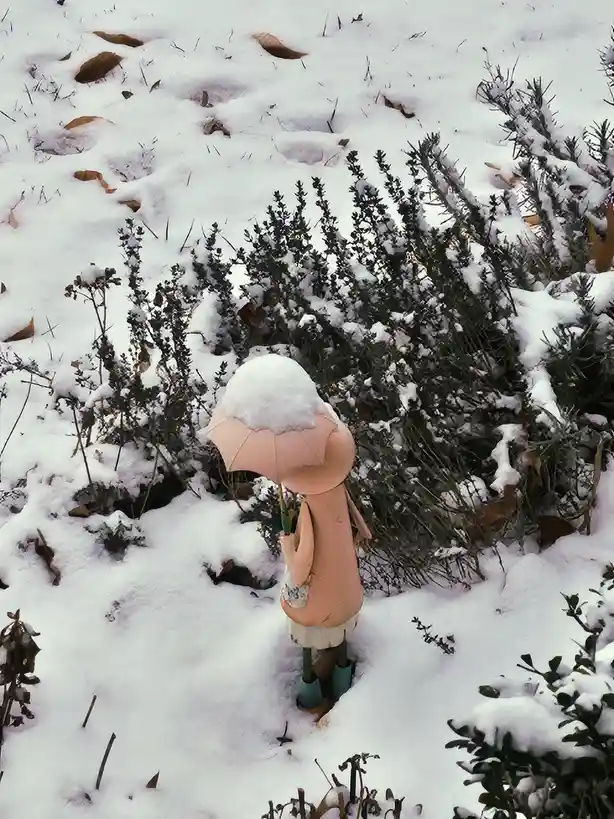 A small, pink, doll-like figurine stands in a snowy garden, holding an umbrella that is completely covered in snow. The…