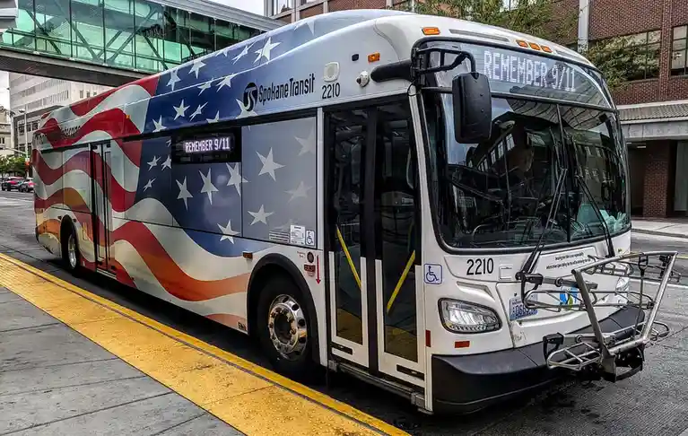 A Spokane Transit bus, decorated with an American flag design, displays the text 'REMEMBER 9/11' on both its front digital…