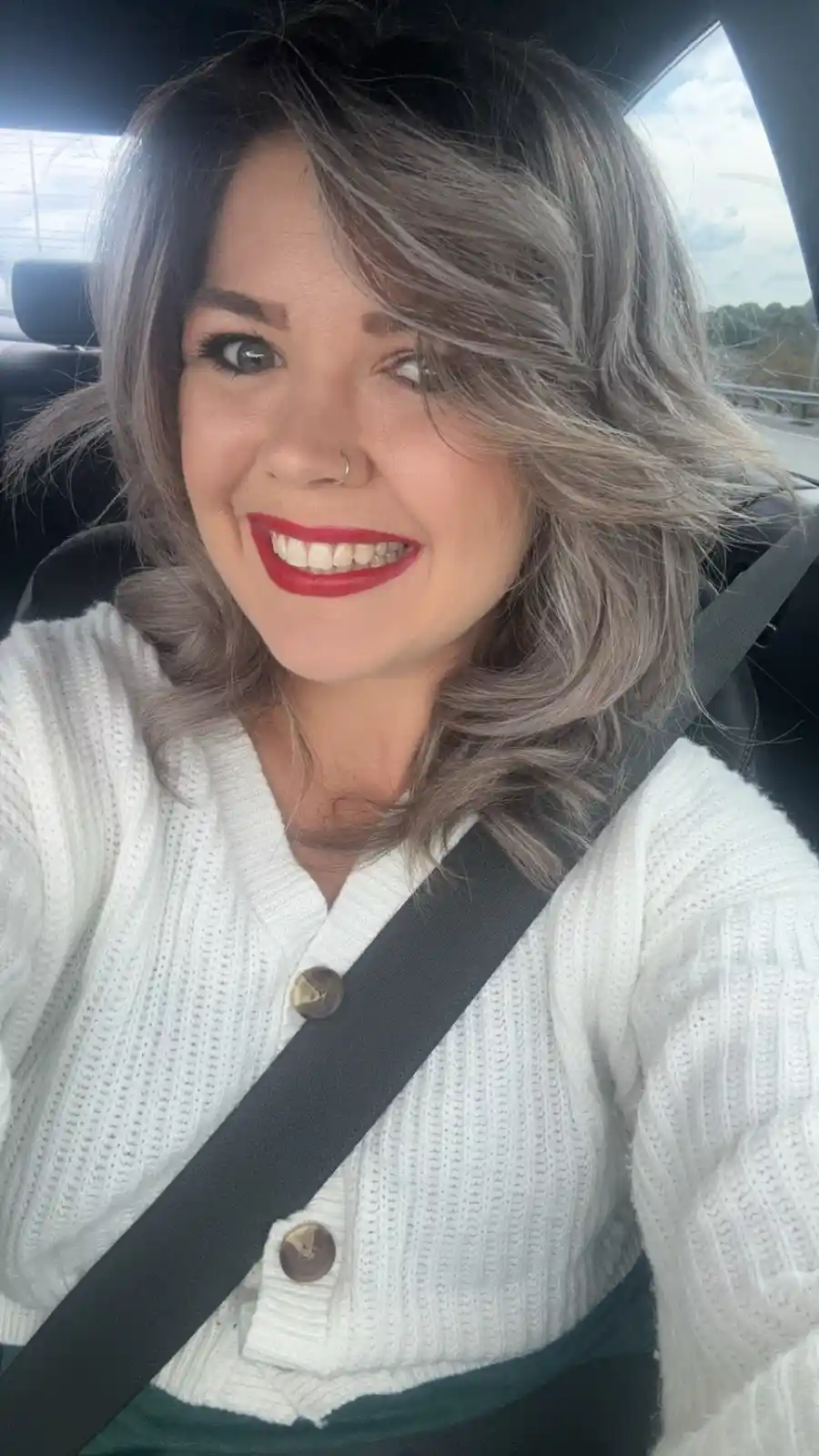 A woman with silver-gray hair and red lipstick smiles at the camera while seated in a car, wearing a white knit cardigan and…