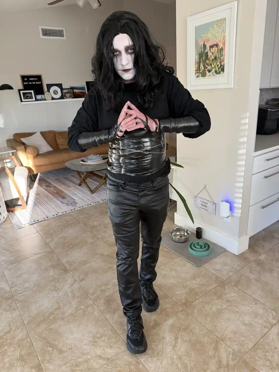 A person dressed in a black wig, white face paint with black tears, and black clothing stands in a living room, their torso…
