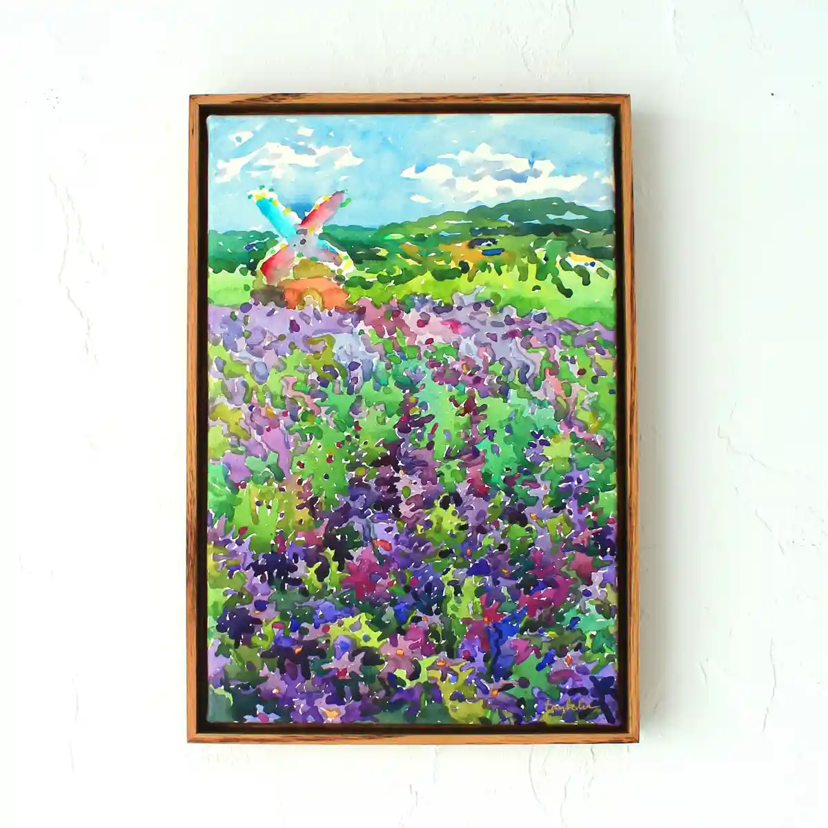 A watercolor painting in a wooden frame hangs on a white wall, depicting a vibrant field of purple and green flowers with a…