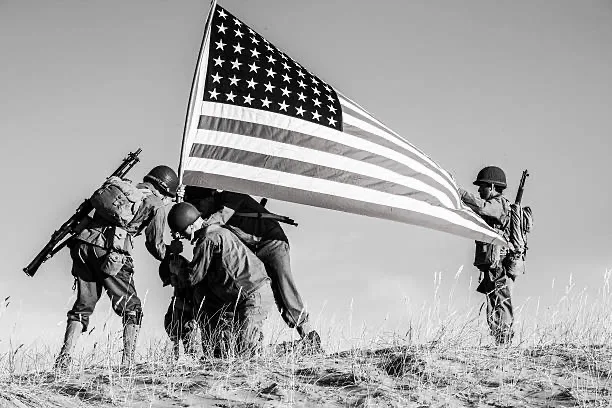four soldiers in military gear are raising a large american flag on a barren hilltop, with one soldier kneeling to secure…