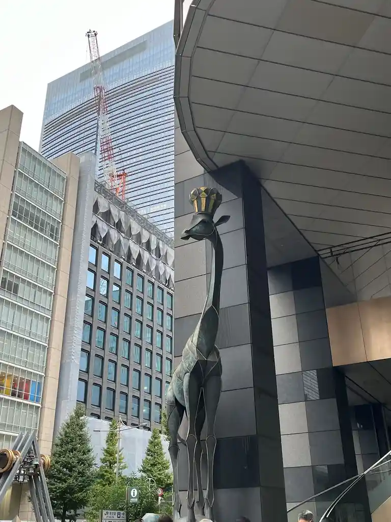 A tall, stylized giraffe statue with a geometric, metallic finish stands in front of a modern building, wearing a golden…
