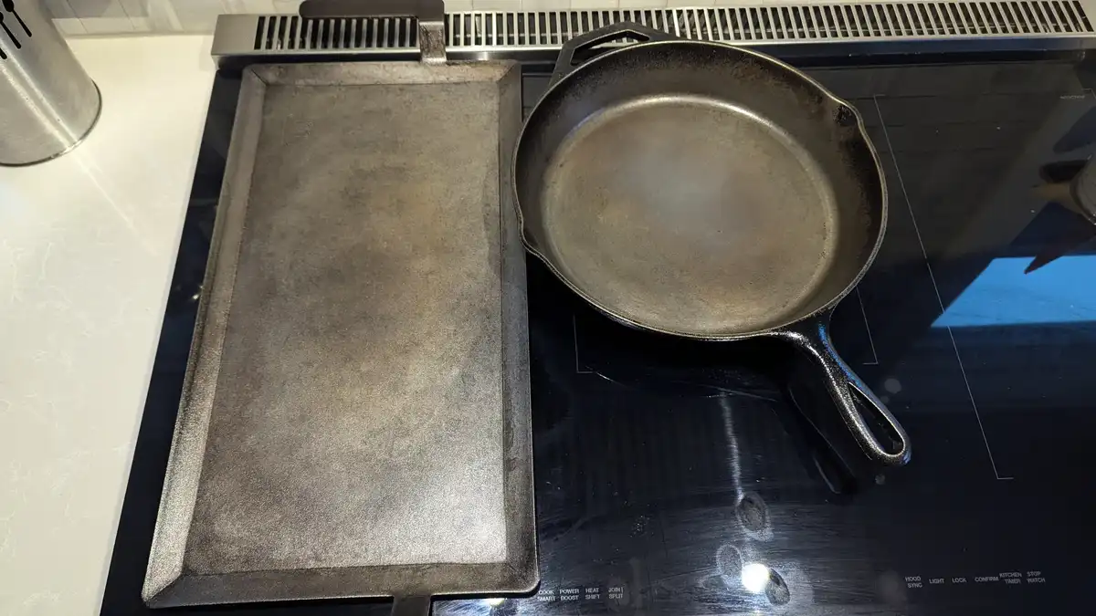 An overhead shot shows two well-used cast iron pans — a rectangular griddle and a round skillet — sitting side-by-side on a…
