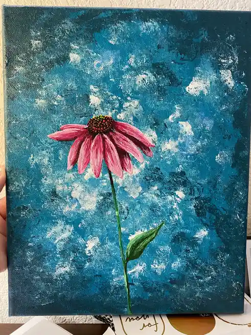 A hand holds a square canvas painting depicting a single pink coneflower with a green stem and leaf against a textured blue…