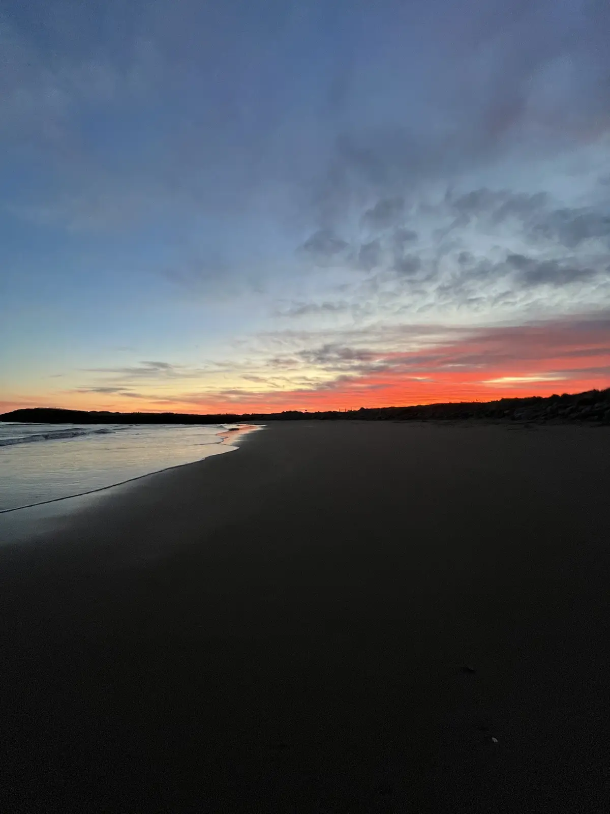 A serene beach scene at sunset, with vibrant orange and pink clouds against a blue sky, waves gently lapping at a dark…