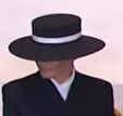 A person wearing a black bowler hat with a white band, their face obscured by the hat's brim, dressed in a dark suit and…